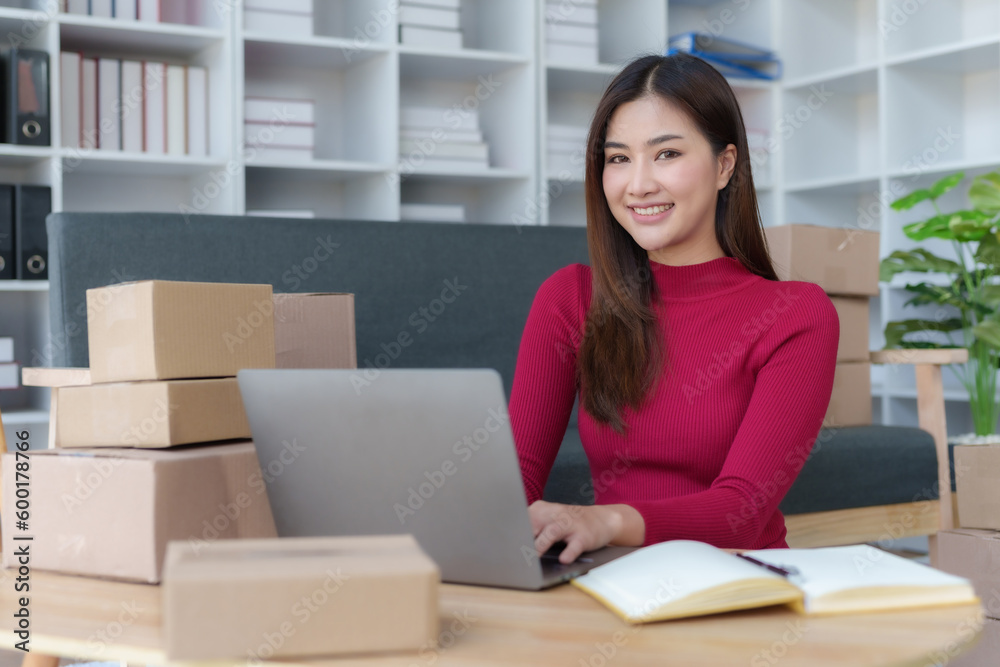 Smiling young Asian business owner woman prepare parcel box and check online orders of product for deliver to customer on laptop computer at home. Shopping Online concept. Startup small business SME. Smiling young Asian business owner woman prepare parcel box and check online orders of product for deliver to customer on laptop computer at home. Shopping Online concept. Startup small business SME.