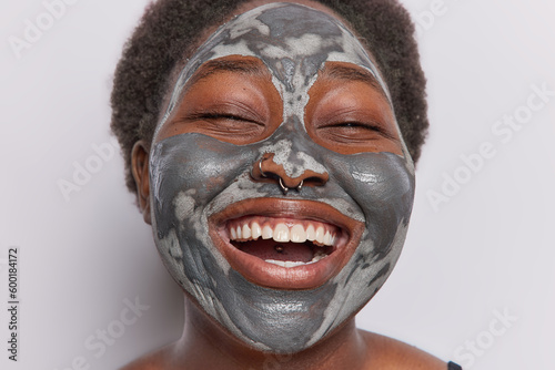 Close up of happy dark haired woman smiles toothily applies clay beauty mask for reducing pores has piercing in nose being very glad as undergoes skin care procedures isolated over white background