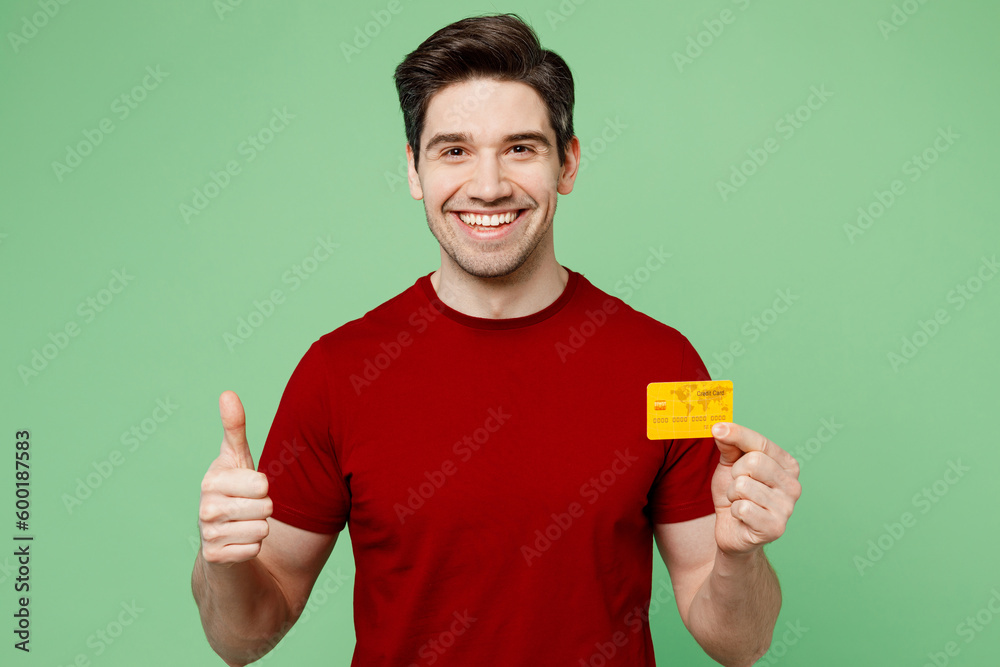 Young smiling happy man he wearing red t-shirt casual clothes hold in hand mock up of credit bank card show thumb up isolated on plain pastel light green background studio portrait. Lifestyle concept. - obrazy, fototapety, plakaty 