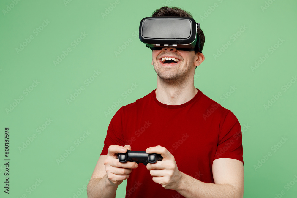 Young excited man wearing red t-shirt casual clothes hold in hand play pc game with joystick console watching in vr headset pc gadget isolated on plain pastel light green background studio portrait. Young excited man wearing red t-shirt casual clothes hold in hand play pc game with joystick console watching in vr headset pc gadget isolated on plain pastel light green background studio portrait.