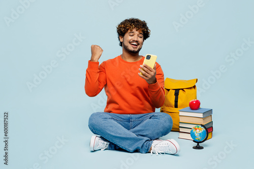 Full body young teen Indian boy student wear casual clothes sit near backpack bag pile of books use mobile phone do winner gesture isolated on plain pastel blue background High school college concept © ViDi Studio