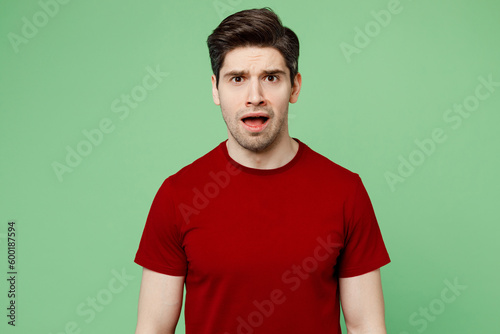 Young shocked astonished sad brunet caucasian man he wears red t-shirt casual clothes look camera with opened mouth isolated on plain pastel light green background studio portrait. Lifestyle concept.