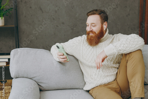 Young smiling ginger man wear casual clothes hold in hand use mobile cell phone sits on grey sofa couch stay at home hotel flat rest relax spend free spare time in living room indoor. Lounge concept.