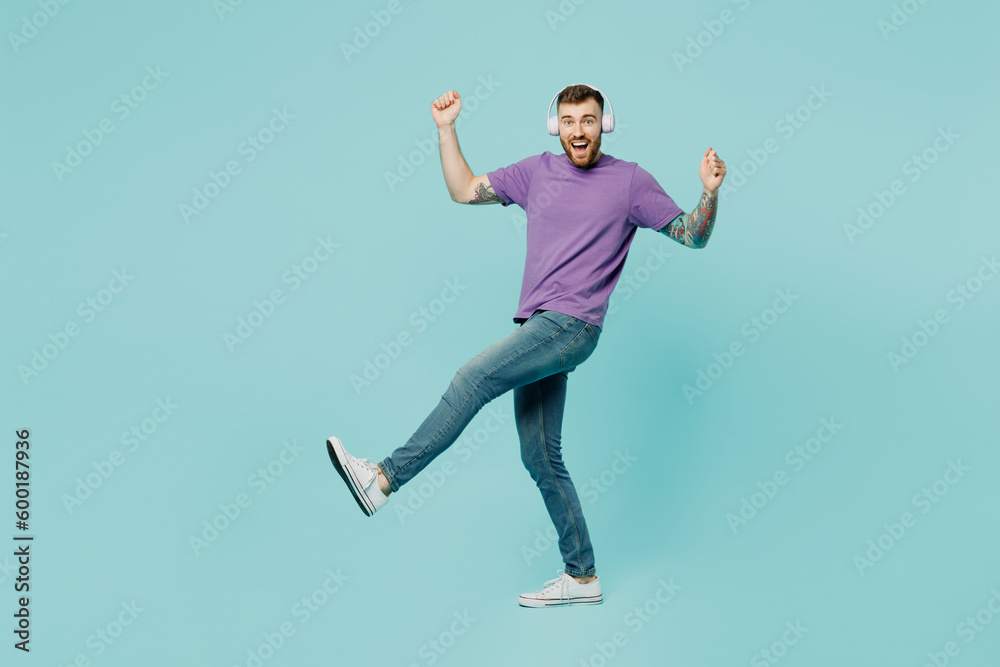 Full body side view cheerful fun young man he wears purple t-shirt headphones listen to music do winner gesture isolated on plain pastel light blue cyan background studio portrait. Lifestyle concept.