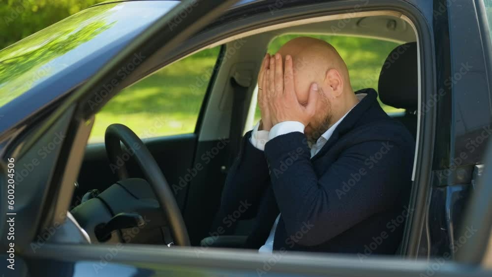 A man in a blue suit is sitting in a car, clutching his head and getting upset. The concept of failure and bad mood