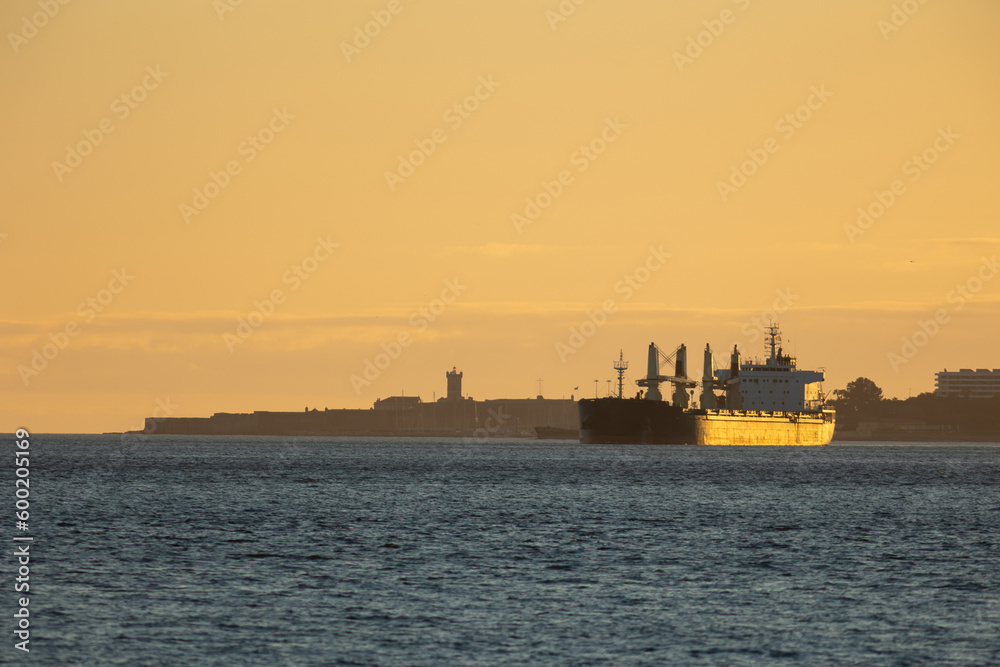 An industrial ship sails down the river at sunset