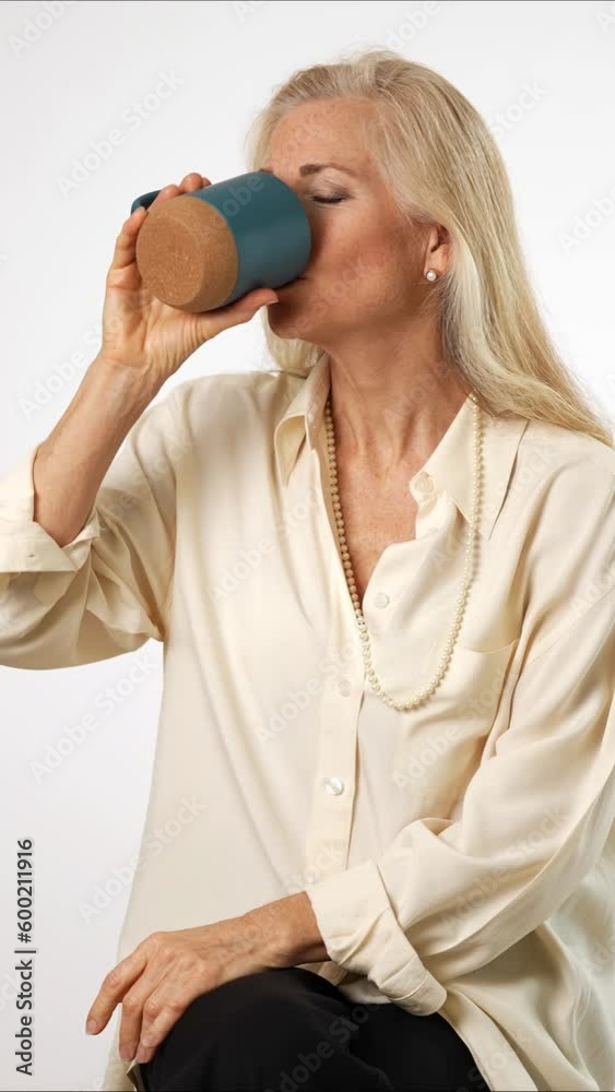 Vertical video of elegant beautiful smiling happy blond mature woman holding cup drinking coffee or tea looking to the side on solid white background