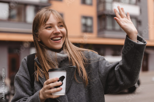 Smiling young woman waving her hand with a friendly cheerful smile to her neighbours. Girl wear grey sweater and coat, hold cup of tea, meeting friends, welcome, nice to meet you. Hi, hello gesture.  photo