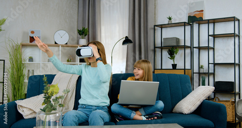 Beautiful positive modern two different ages girls spending joint leisure at playing virtual games using laptop and augmented reality goggles,front view