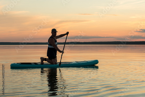 A man on his knees on a SUP board with an oar at sunset swims in the water of the lake against a pink sky reflection in the water. © finist_4