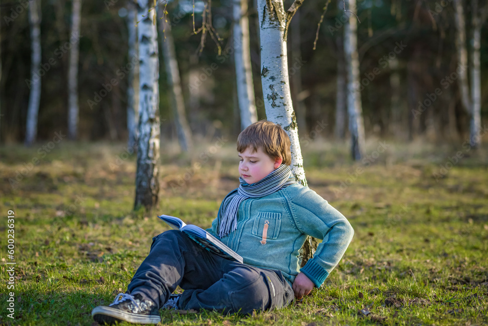 A handsome boy in a scarf is in a spring park, sitting under a birch tree, reading a book and enjoying his dreams. A handsome boy in a scarf is in a spring park, sitting under a birch tree, reading a book and enjoying his dreams.