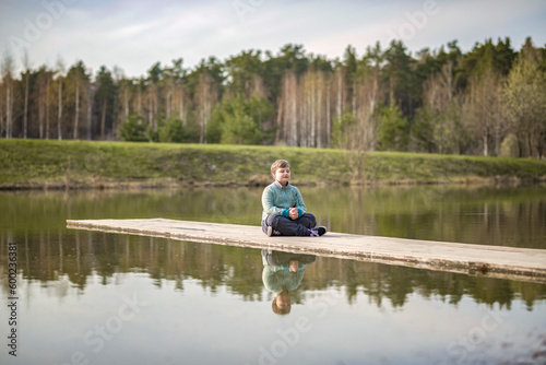 A boy is sitting on a bridge in a green park. The path is a bridge over the lake.