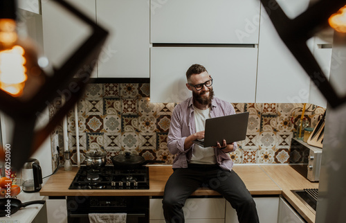 Charming male freelancer with a beard in casual clothes works remotely at a laptop from home sitting in the kitchen  communicating with a client via the Internet.