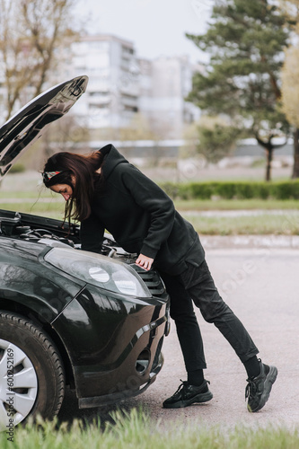 A young woman tries to repair a broken black car by making an inspection into the engine under the hood. Photography, maintenance concept.