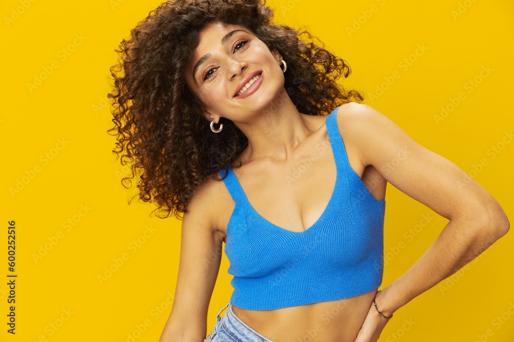 Woman with curly afro hair in a blue t-shirt on. yellow background signs with her hands, look into the camera, smile with teeth and happiness, copy space Woman with curly afro hair in a blue t-shirt on. yellow background signs with her hands, look into the camera, smile with teeth and happiness, copy space