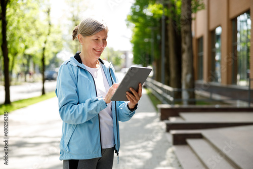 Close View Of The Sporty Mature Runner Woman Standing In The Street And Swipes On The Tablet Screen or Texting. Sport And Healthy Lifestyle Concept