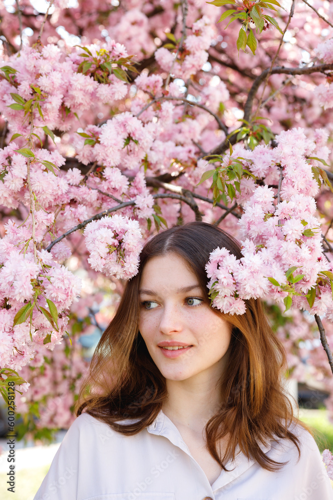 Side View of the Attractive Girl with Sakura Flowers in her Hair Posing Against Blooming Tree Outside. Dark Haired Woman Looking Away. Femininity and Natural Beauty Concept Side View of the Attractive Girl with Sakura Flowers in her Hair Posing Against Blooming Tree Outside. Dark Haired Woman Looking Away. Femininity and Natural Beauty Concept