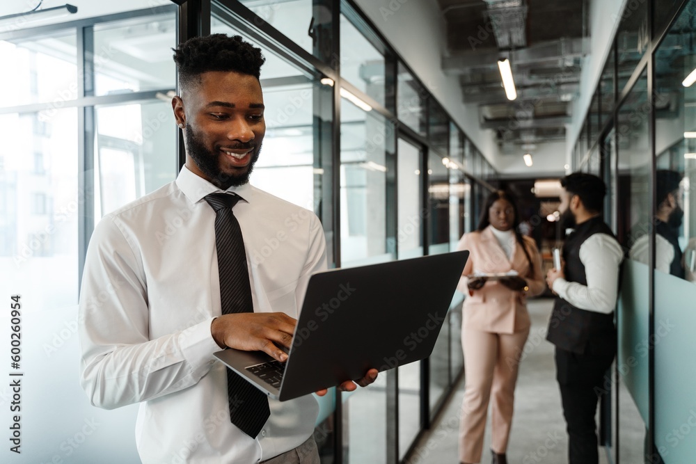 Young african american man in business suit holding laptop while standing at office corridor Young african american man in business suit holding laptop while standing at office corridor