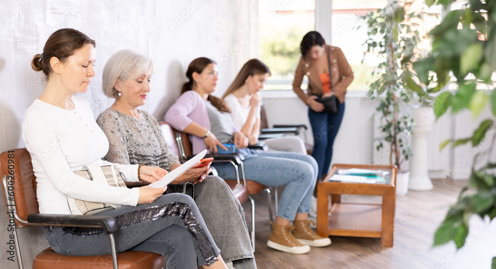 Fototapeta premium Adult woman in casual clothes reads text of document while sitting on chair in waiting room waiting for her turn