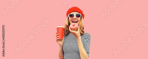 Portrait of happy cheerful young french woman eating sweet donut with cup of coffee or juice in heart shaped sunglasses and beret hat on pink background
