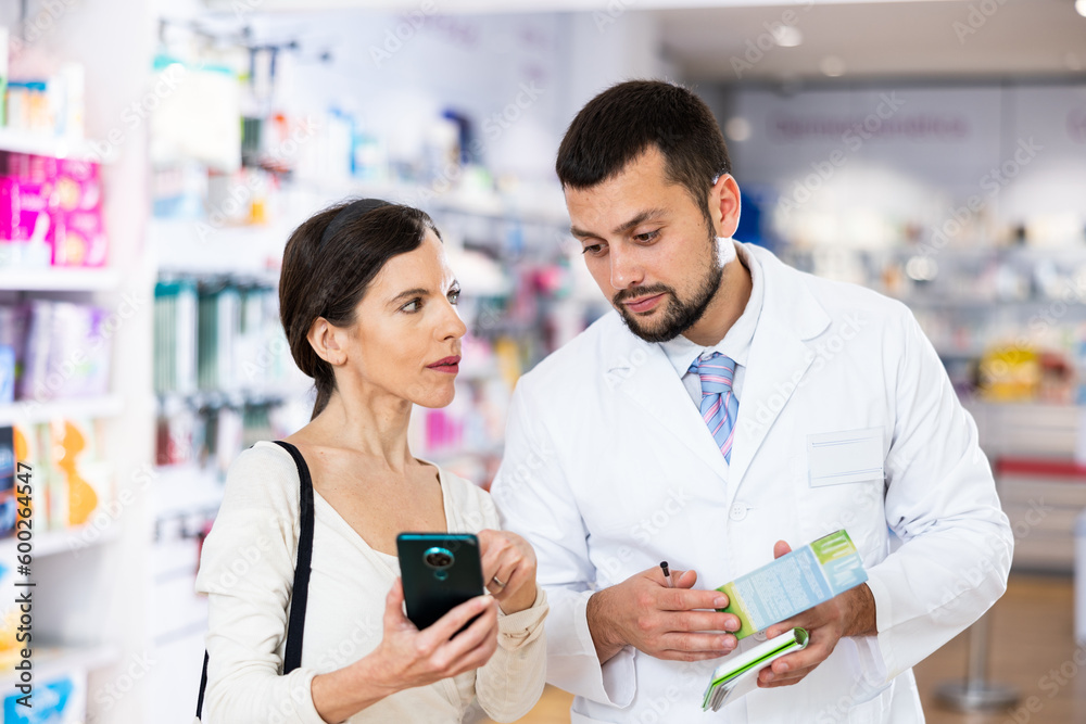 Fototapeta premium Friendly man pharmacist helping young adult woman with electronic prescription in her phone choosing medical supplies at drugstore