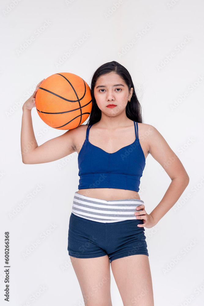 A sporty and athletic young woman posing while holding basketball between her hand and shoulder. Isolated on a white background.