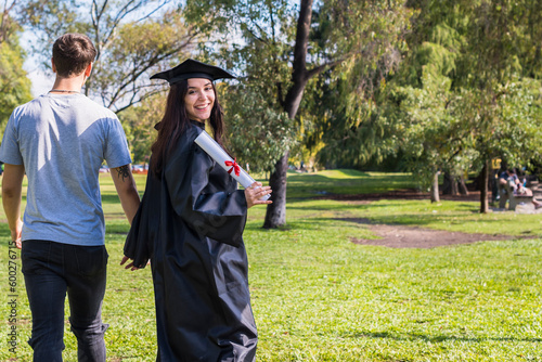 Happy graduated girl walking whitle holding hands with her boyfriend while she turns arround to show her diploma. Copy space.