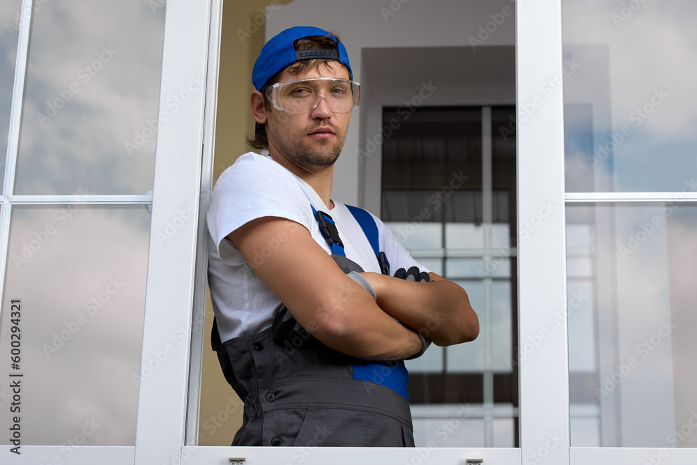 Brutal man with languid look in construction clothes and protective glasses stands in opening window with hands folded on chest, looking into camera. Portrait young serious installer while working