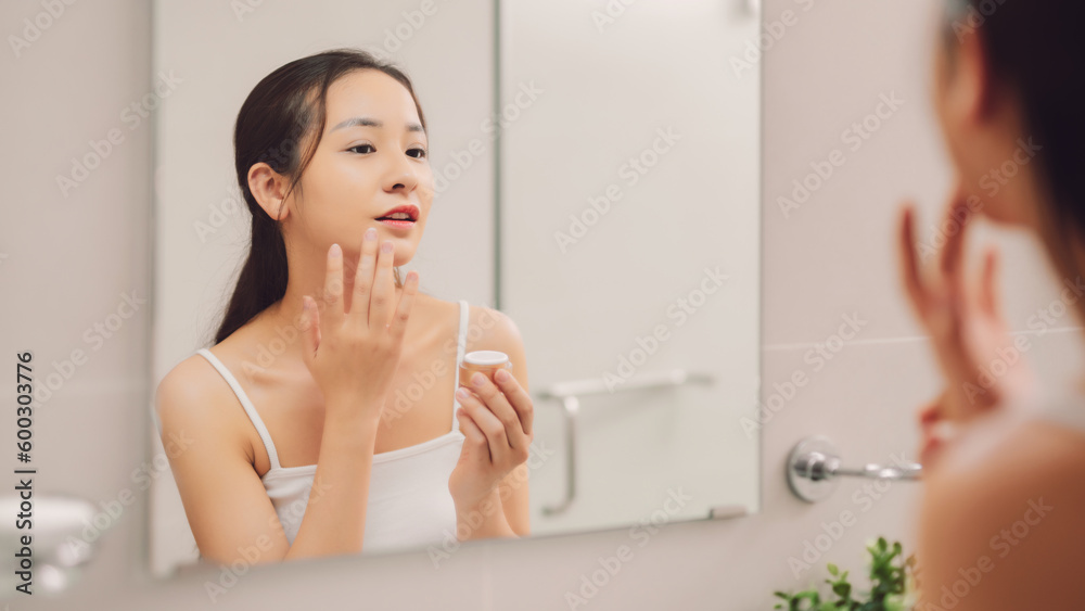 Woman caring of her beautiful skin on the face standing near mirror in the bathroom. Young woman applying moisturizer on her face. Smiling girl holding little jar of skin cream and applying lotion. Woman caring of her beautiful skin on the face standing near mirror in the bathroom. Young woman applying moisturizer on her face. Smiling girl holding little jar of skin cream and applying lotion.