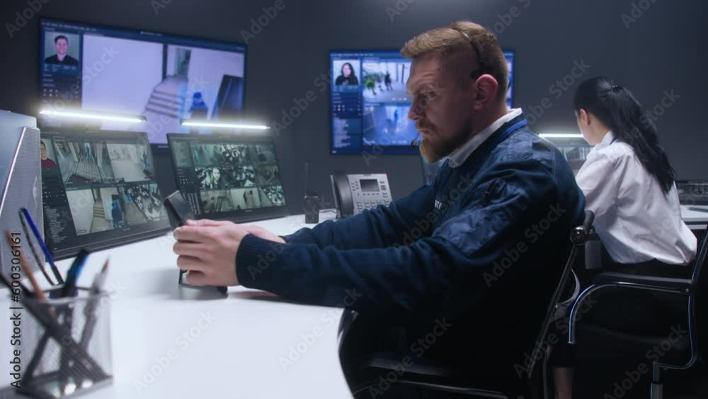 Male security officer uses tablet and controls CCTV cameras with AI facial recognition in police surveillance center. PC monitors and big digital screens showing security cameras footage. Monitoring. Male security officer uses tablet and controls CCTV cameras with AI facial recognition in police surveillance center. PC monitors and big digital screens showing security cameras footage. Monitoring.