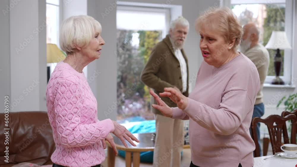 Caucasian senior women arguing as blurred husbands ignoring wives standing at background talking. Side view unsatisfied old friends quarreling in living room indoors Caucasian senior women arguing as blurred husbands ignoring wives standing at background talking. Side view unsatisfied old friends quarreling in living room indoors