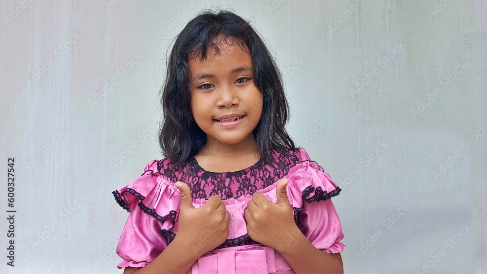 Close up portrait little girl smiling wearing pink dress giving thumb up Close up portrait little girl smiling wearing pink dress giving thumb up