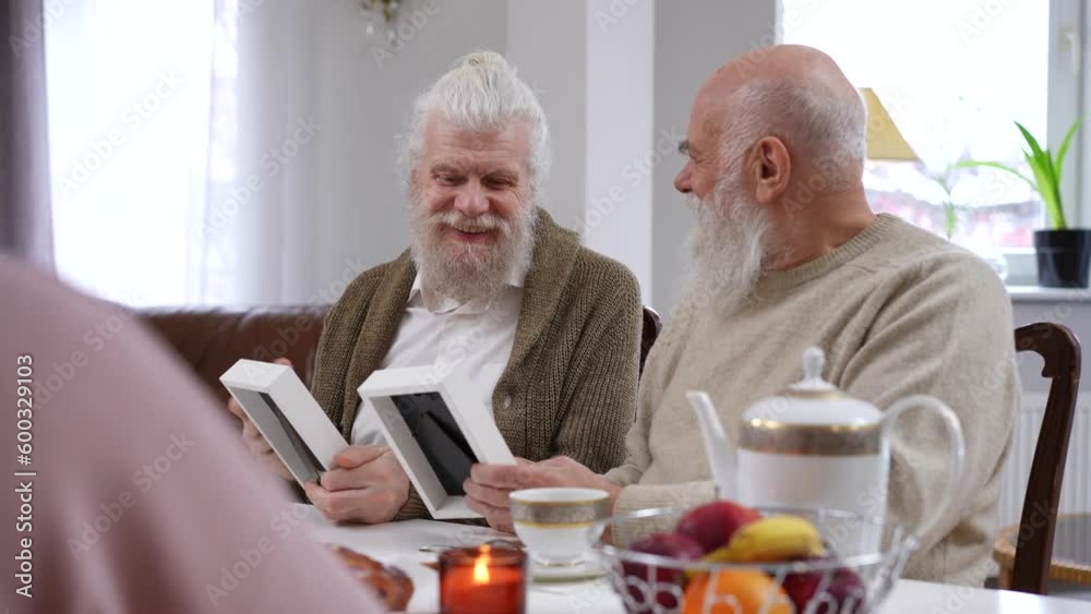 Joyful bearded senior men talking watching old photographs sitting at table in living room. Positive cheerful Caucasian retirees friends chatting enjoying nostalgia Joyful bearded senior men talking watching old photographs sitting at table in living room. Positive cheerful Caucasian retirees friends chatting enjoying nostalgia