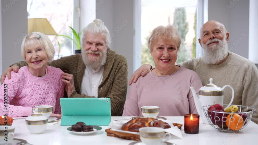 Medium shot positive Caucasian old couples hugging smiling and looking at camera. Front view portrait of senior men and women posing sitting at table in living room indoors Medium shot positive Caucasian old couples hugging smiling and looking at camera. Front view portrait of senior men and women posing sitting at table in living room indoors