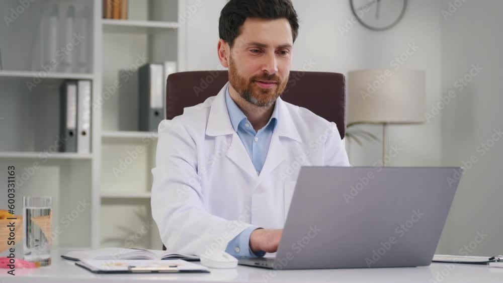 Positive man is smiling. By the table with laptop. Doctor in formal clothes is working in the cabinet. Positive man is smiling. By the table with laptop. Doctor in formal clothes is working in the cabinet.
