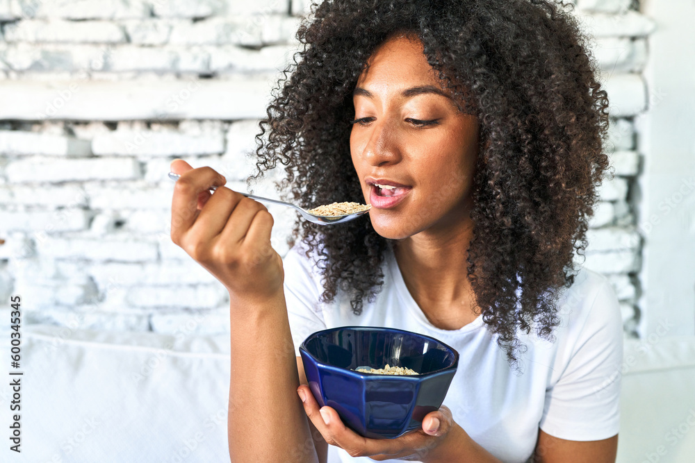African-American woman savors a comforting bowl of oatmeal on the sofa, starting her day with nourishment. - obrazy, fototapety, plakaty 