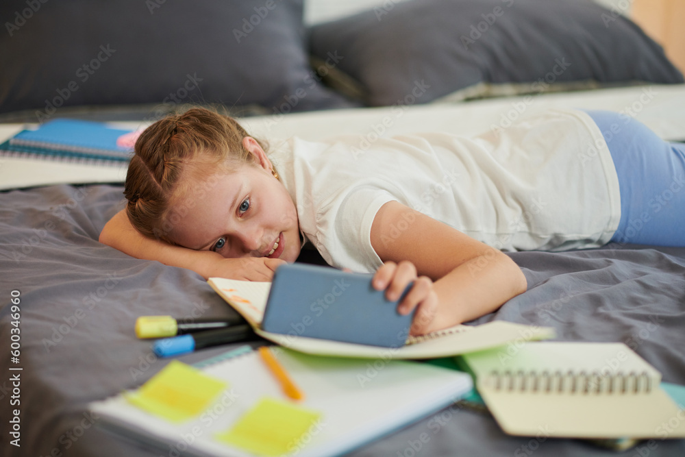 Smiling girl resting on bed and watching entertaining videos on smartphone after doing homework