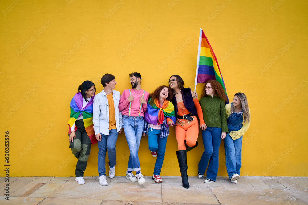 Obraz premium Portrait of group cheerful young friends standing on wall yellow. LGBT community smiling people on gay pride day enjoying together and showing rainbow flags. Generation z and parade party homosexual.