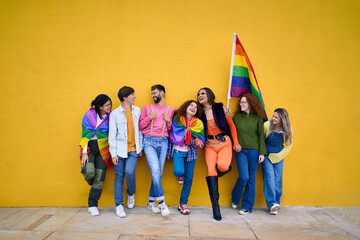 Portrait of group cheerful young friends standing on wall yellow. LGBT community smiling people on gay pride day enjoying together and showing rainbow flags. Generation z and parade party homosexual.