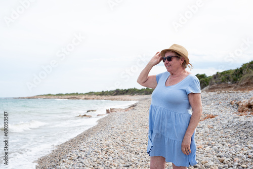 Smiling senior woman holding her sunhat and looking at Mediterranean sea on a pebble beach.