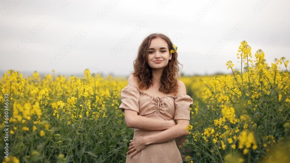 Portrait of a smiling woman standing in a flower rape field outdoors. A coquettish girl with curly hair smiles sweetly. Slow motion. Teenage girl in a blooming field on a summer day. Portrait of a smiling woman standing in a flower rape field outdoors. A coquettish girl with curly hair smiles sweetly. Slow motion. Teenage girl in a blooming field on a summer day.
