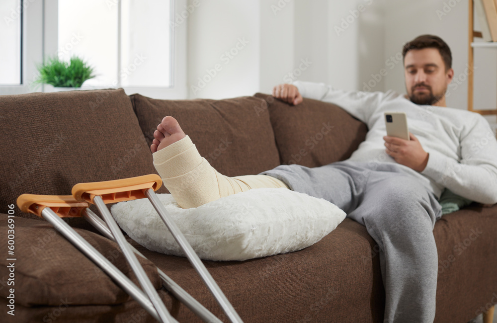 Young man with crutches is lying on the couch at home with broken leg