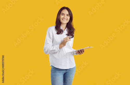 Portrait of happy young female secretary, office manager or business assistant with clipboard. Pretty woman in white shirt standing isolated on yellow background, holding clipboard and pen and smiling