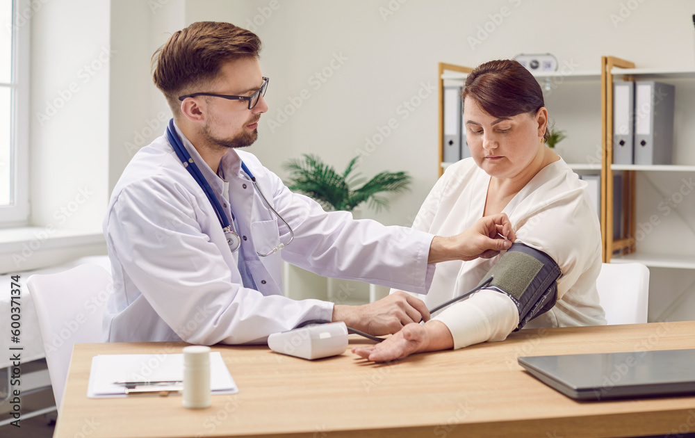 Male doctor working with female patient who has hypertension. Young man physician in white coat sitting at desk with fat, obese woman and using sphygmomanometer to measure her arterial blood pressure Male doctor working with female patient who has hypertension. Young man physician in white coat sitting at desk with fat, obese woman and using sphygmomanometer to measure her arterial blood pressure