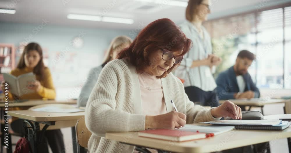 Elderly Woman Acquiring New Academic Skills in Classroom with Other People. Adult Class Writing Down Notes in Notebook, Taking a Test or Exam. Teacher Walking Between Desks Elderly Woman Acquiring New Academic Skills in Classroom with Other People. Adult Class Writing Down Notes in Notebook, Taking a Test or Exam. Teacher Walking Between Desks