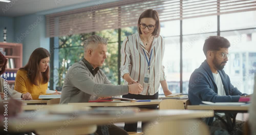 Helpful Young Teacher Talking with an Adult Man, Helping to Find a Solution for a Test Course Assignment. Multiethnic Women and Men of Different Age Acquiring New Academic Skills in Classroom Helpful Young Teacher Talking with an Adult Man, Helping to Find a Solution for a Test Course Assignment. Multiethnic Women and Men of Different Age Acquiring New Academic Skills in Classroom