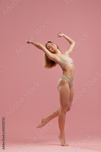 Portrait with beautiful slim girl, model wearing beige seamless underwear posing on fingertips over pink studio background. Beauty and wellness