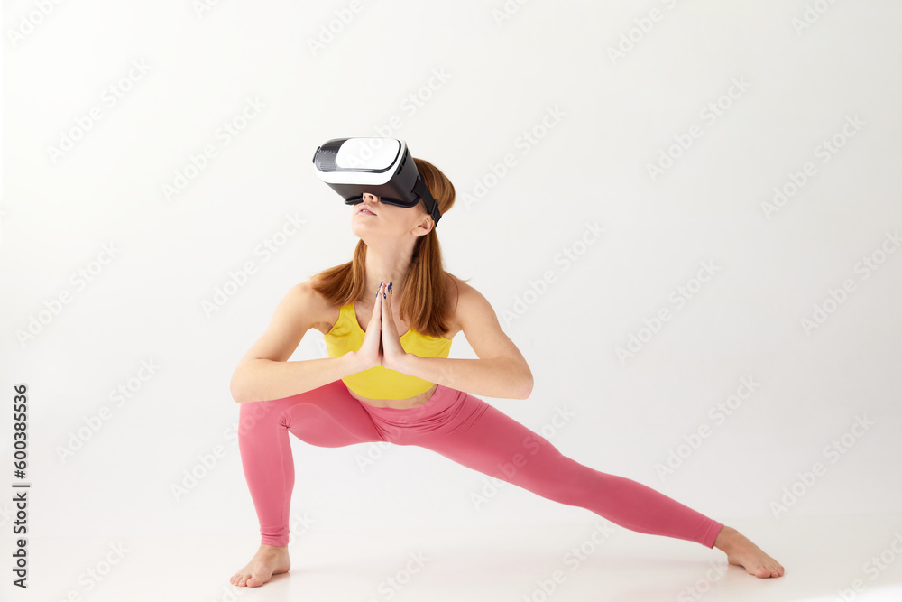 Fototapeta premium Portrait with young girl wearing sportswear and VR glasses doing yoga exercise over white background. Virtual sport training