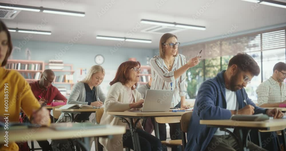 Young Female Teacher Giving a Lecture During an Adult Education Course in School, Having a Conversation with a Older Female with Laptop. Diverse Mature Students Doing Textbook Exercises in Classroom