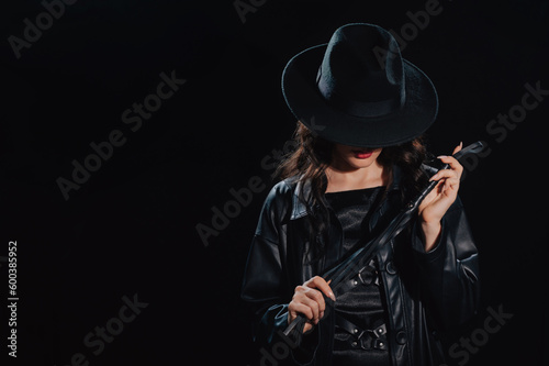 Sexy female dominant mistress with whip for BDSM sex with submission and domination in a leather raincoat and hat in noir style on dark background